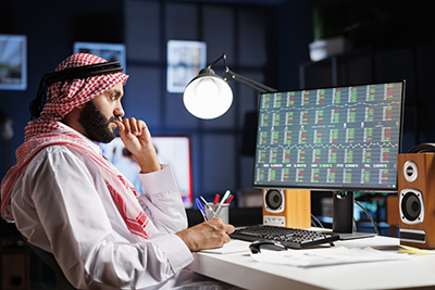 Diligent Muslim man working at a desk with a computer, checking financial reports and taking notes. Focused Arab guy using desktop pc, working with documents at the home office.